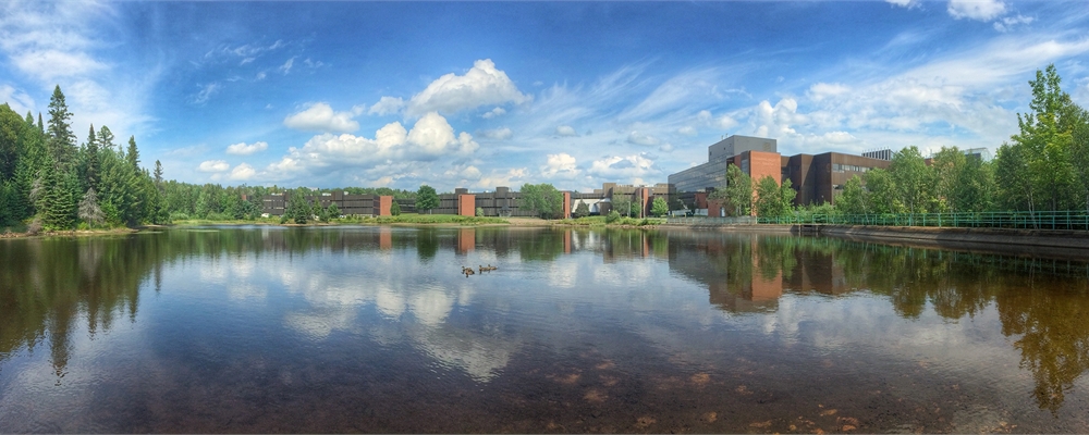 Nipissing University Pond with many trees and blue cloudy skies.